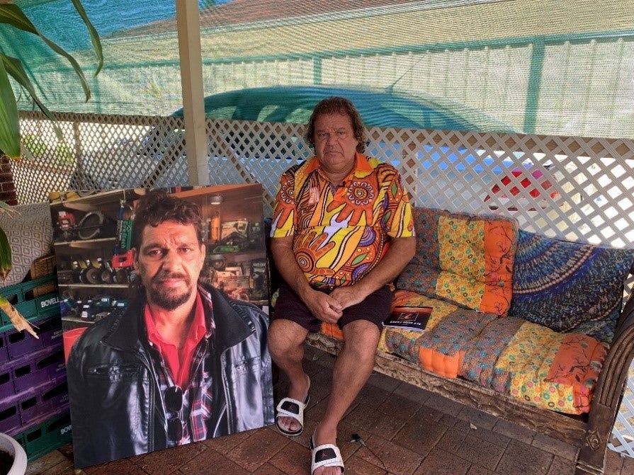 A man sits on a couch next to a large photo of his brother