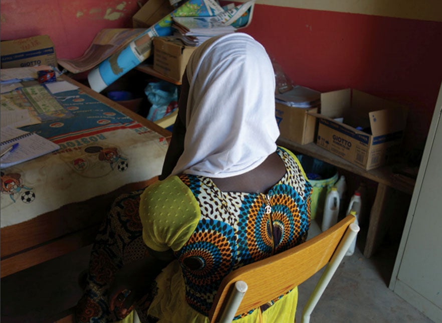 A girl sits in a chair, with her back facing the camera, in a colorful dress and a white headscarf.