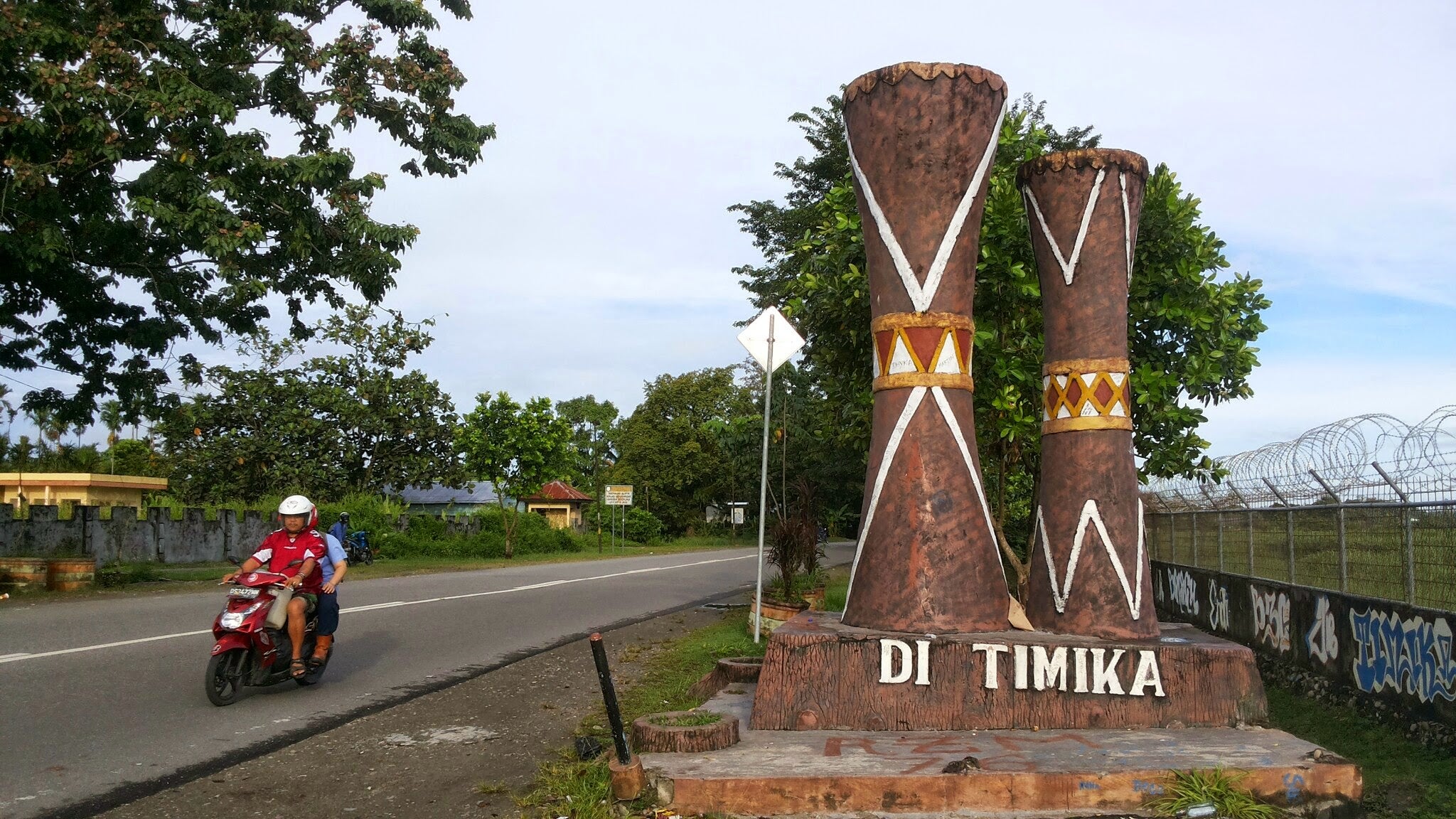 People on a motorcycle ride past a statue of two traditional Papuan wood drums