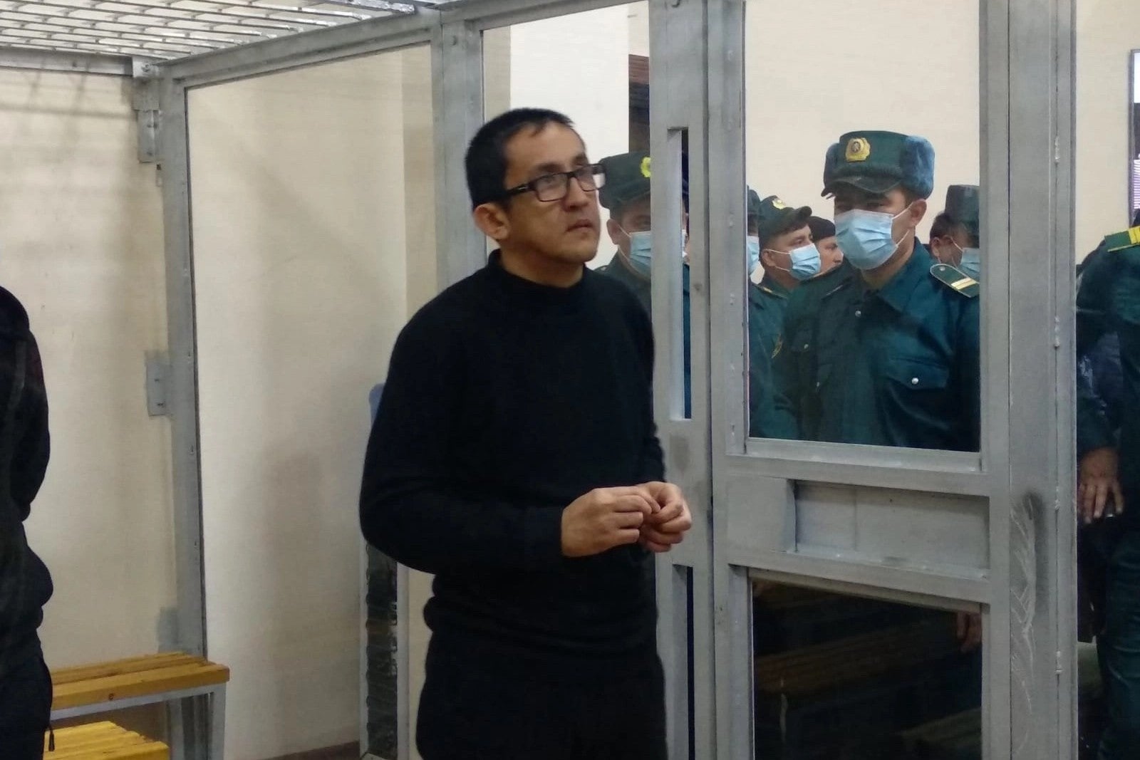 A man stands inside a glass defendants cage in a courtroom