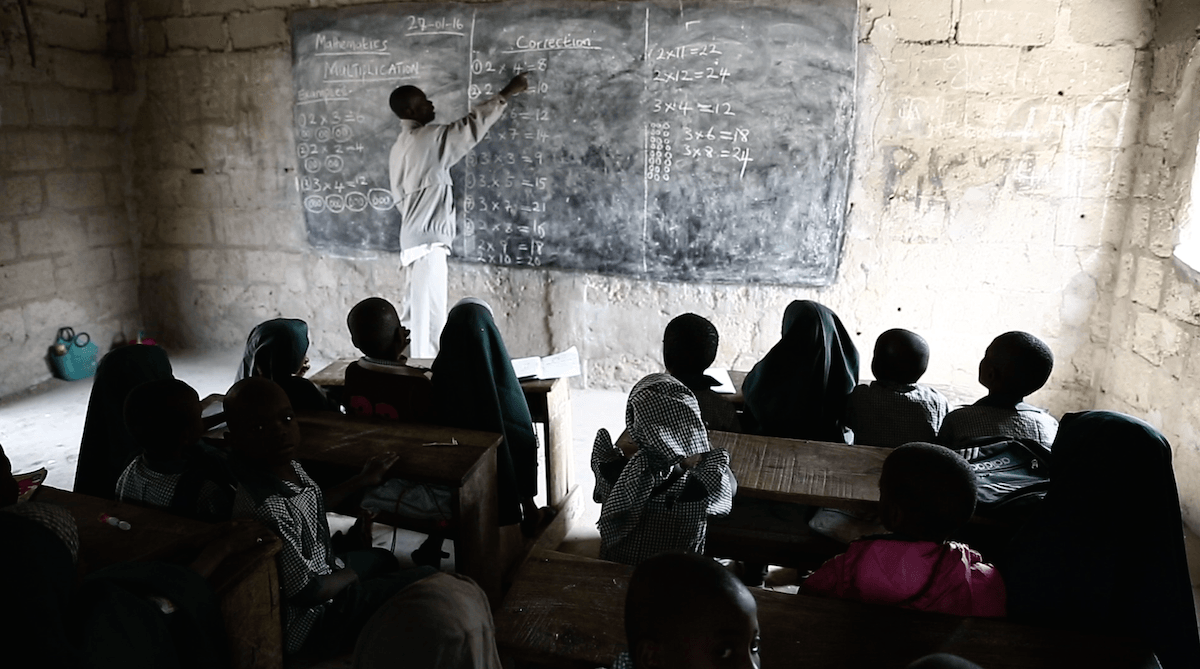 A picture of a teacher writing on a black board while students pay attention.