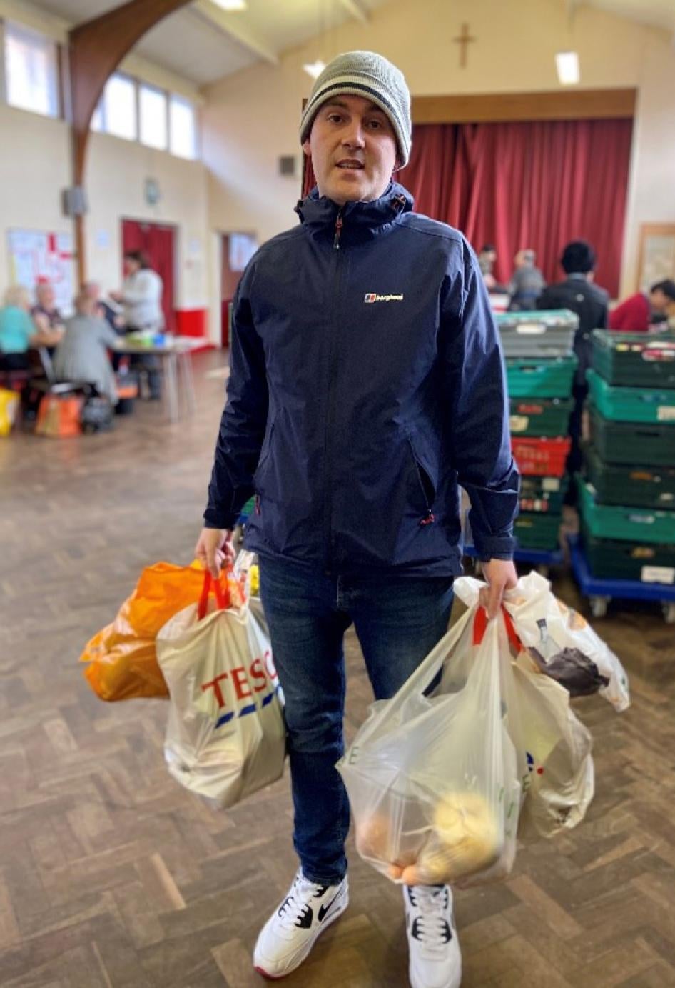 A man in a beanie and jacket holds bags of groceries 