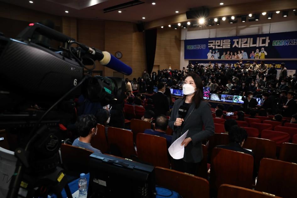 A woman in a face mask holds a microphone in front of a video camera