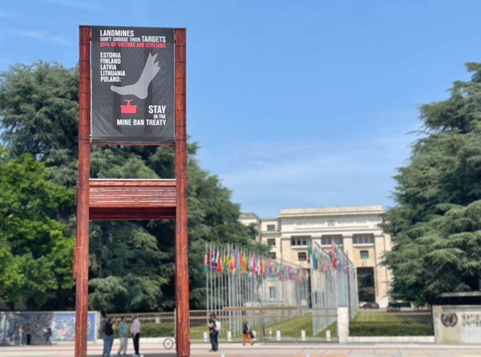 A banner hangs on the Broken Chair sculpture stating "Landmines don't choose their targets. 85% of victims are civilians. Estonia, Finland, Latvia, Lithuania, and Poland stay in the Mine Ban Treaty."