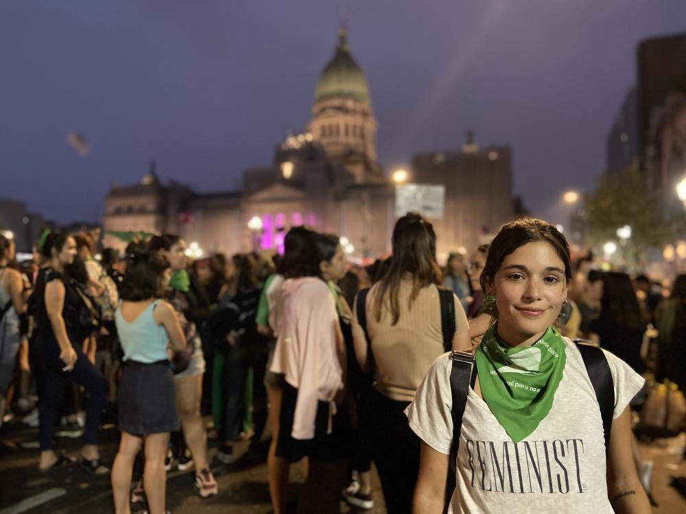A woman wearing a t-shirt that says "Feminist" on it poses amidst a rally
