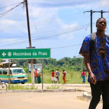 A man walks down a street with a sign for the town "Mocimboa da Praia" behind him