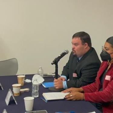 A man seated at a table speaks into a microphone